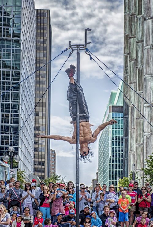 A very fit, shritless male hangs by his feet from an erect pole, surrounded by onlookers in a downtown street.